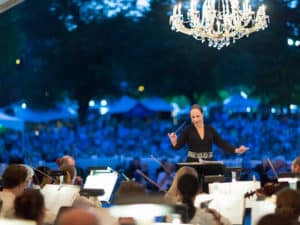 Conductor in an elegant evening gown directs a symphony at dusk, with a chandelier overhead and a large outdoor crowd seated behind her.