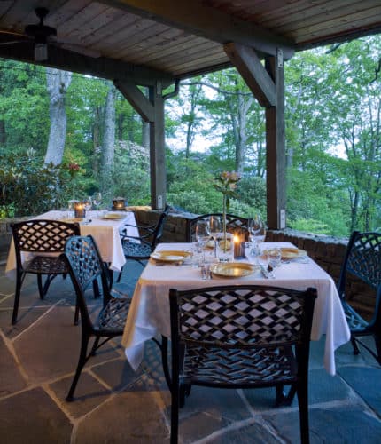 Dining tables set for diners in a wooded setting, with tablecloths and candlelight.