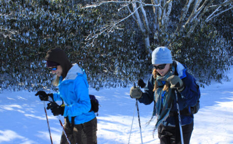Two women in winter gear hike with poles in the snow.