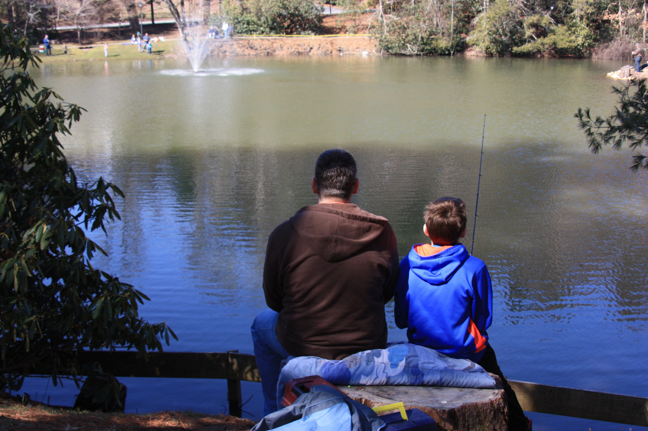 A father a son sit next to each other with their backs to the camera, facing a pond with a fountain. The boy is holding a fishing pole.
