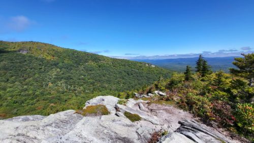 A rock cliff with a view of a slope to the left and layers of blue mountains in the distance to the right.