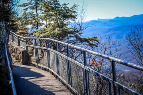 A paved trail with a guardrail overlooks a distant view of blue mountains.