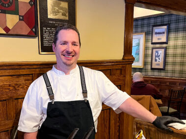 A young man in a chef coat and black apron smiles for the camera