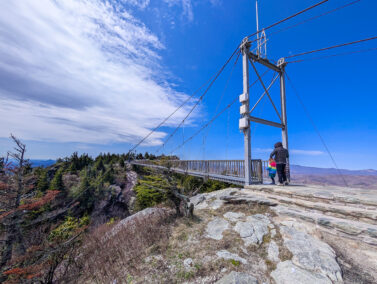 A father and daughter at the edge of a footbridge- the Grandfather Mountain Mile High Swinging Bridge- under bright, blue sky.