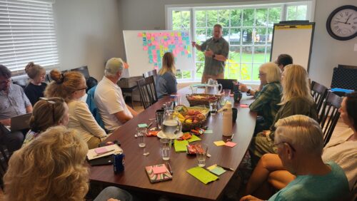 A person stands in front of a room of people seated at a long table, leading a discussion in front of a whiteboard.