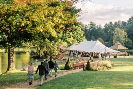 A group of people walk toward an outdoor event tent on a wide lawn surrounded by trees.