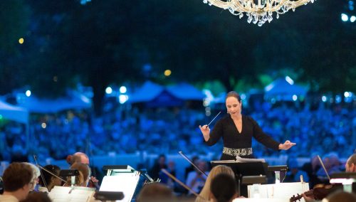 Conductor in an elegant evening gown directs a symphony at dusk, with a chandelier overhead and a large outdoor crowd seated behind her.